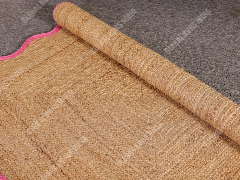 May include: A rolled-up, natural-toned jute rug with a pink scalloped border. The rug has a woven texture and a geometric pattern. The rolled portion reveals the rug's thickness and construction. The rug is on a gray surface.