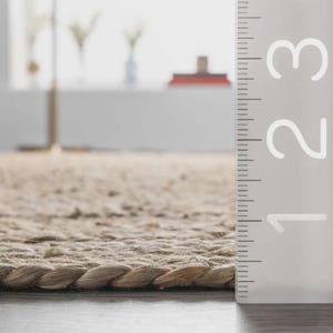 May include: Close-up of a beige jute rug with a braided edge. The rug's texture is visible, and a ruler is positioned vertically along the right side, showing measurements up to 3 inches.