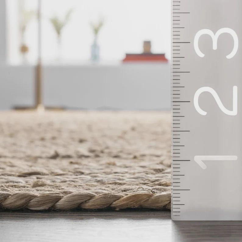 May include: Close-up of a beige jute rug with a braided edge. The rug's texture is visible, and a ruler is positioned vertically along the right side, showing measurements up to 3 inches.