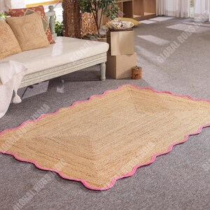 May include: A rectangular jute rug with a scalloped pink border. The rug is made of natural fibers and has a light brown color. It is placed on a gray carpeted floor, near a white couch with beige pillows.