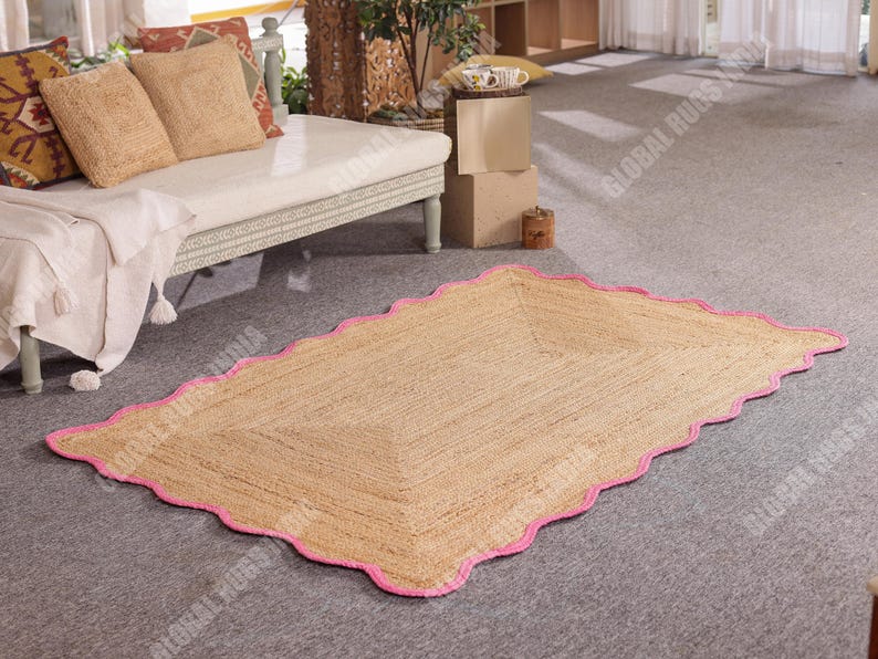 May include: A rectangular jute rug with a scalloped pink border. The rug is made of natural fibers and has a light brown color. It is placed on a gray carpeted floor, near a white couch with beige pillows.