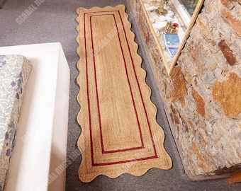 Puede incluir: Una alfombra rectangular de yute con borde festoneado y borde rojo. La alfombra es de color beige natural y está colocada sobre una superficie gris. La alfombra mide aproximadamente 152 cm de largo.