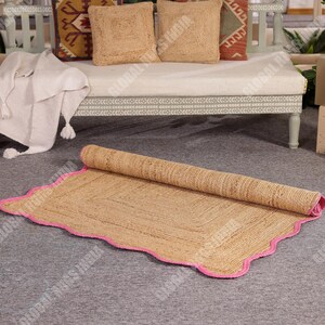 May include: A rolled-up, rectangular jute rug with a pink scalloped border. The rug is a natural tan color and sits on a gray surface. The rug is partially unrolled, revealing its woven texture. The background includes a bench with pillows and a blanket.