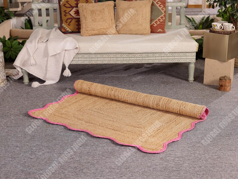 May include: A rolled-up, rectangular jute rug with a pink scalloped border. The rug is a natural tan color and sits on a gray surface. The rug is partially unrolled, revealing its woven texture. The background includes a bench with pillows and a blanket.