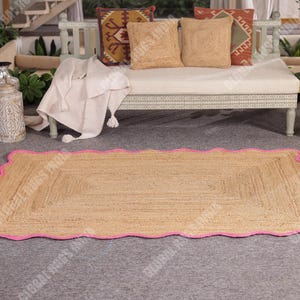 Puede incluir: Una alfombra rectangular de yute con un borde rosa festoneado. La alfombra es de color marr&oacute;n claro y tiene una textura tejida. La alfombra est&aacute; colocada sobre una superficie gris, con un banco y cojines en el fondo.