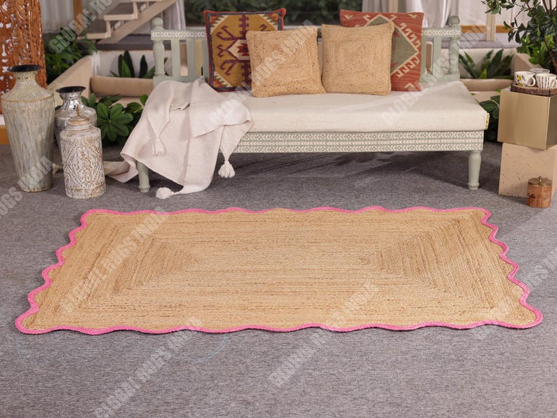 Puede incluir: Una alfombra rectangular de yute con un borde rosa festoneado. La alfombra es de color marr&oacute;n claro y tiene una textura tejida. La alfombra est&aacute; colocada sobre una superficie gris, con un banco y cojines en el fondo.