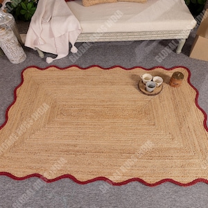 Puede incluir: Una alfombra rectangular de yute con un borde rojo festoneado. La alfombra tiene un color beige natural y una textura tejida. Peque&ntilde;as tazas y un posavasos de madera est&aacute;n sobre la alfombra. Esta alfombra es adecuada para salones y dormitorios.