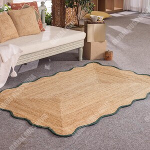 Puede incluir: Una alfombra rectangular de yute con borde festoneado y borde verde. La alfombra es de color marr&oacute;n claro y tiene una textura tejida. La alfombra est&aacute; colocada sobre un suelo alfombrado gris, cerca de un div&aacute;n blanco con cojines.