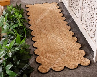 May include: A rectangular jute rug with a scalloped black border. The rug has a natural tan color and a woven texture. The rug is placed on a gray surface near a white decorative cabinet and green plants.