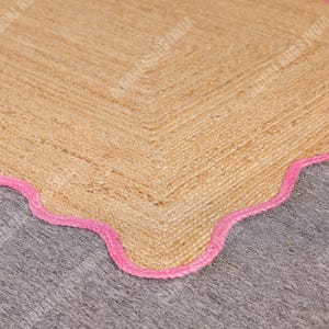 May include: A natural jute rug with a scalloped edge in bright pink. The rug has a woven texture and a neutral beige color, with a decorative border. This rug is suitable for home decor.