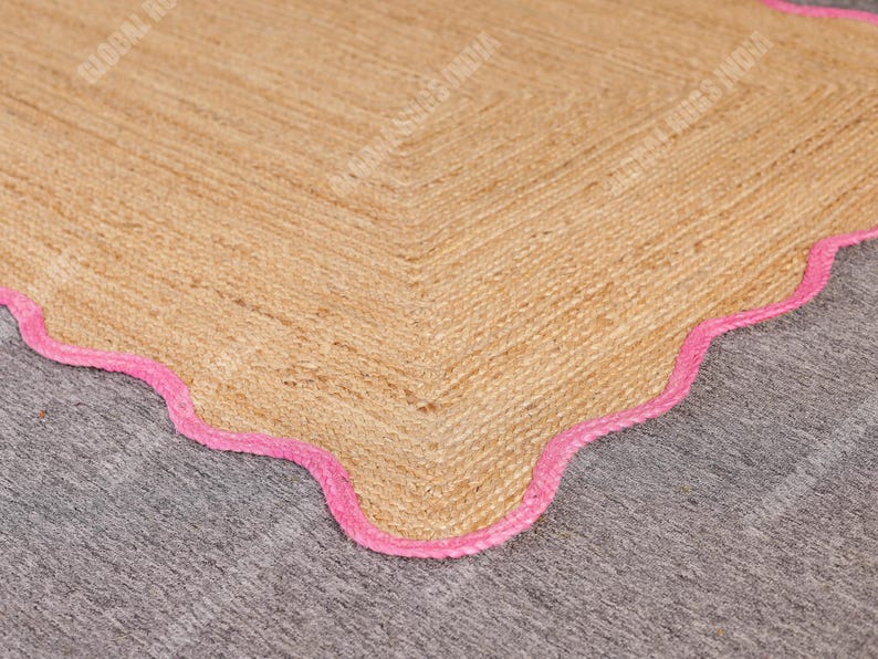 May include: A natural jute rug with a scalloped edge in bright pink. The rug has a woven texture and a neutral beige color, with a decorative border. This rug is suitable for home decor.