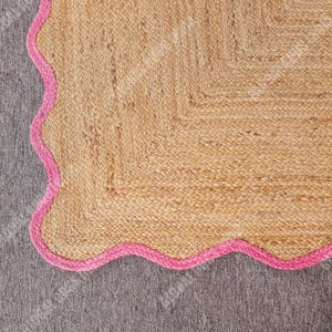May include: A natural fiber rug with a scalloped pink border. The rug is made of woven jute fibers in a light tan color. The pink border adds a pop of color and a decorative touch. The rug is placed on a gray surface.