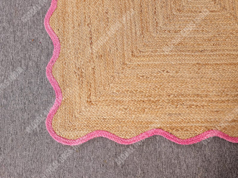 May include: A natural fiber rug with a scalloped pink border. The rug is made of woven jute fibers in a light tan color. The pink border adds a pop of color and a decorative touch. The rug is placed on a gray surface.