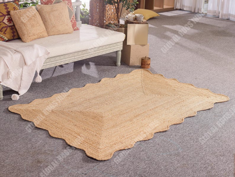 Puede incluir: Una alfombra rectangular de yute con borde festoneado. La alfombra es de color beige natural y tiene una textura tejida. Se coloca sobre un suelo alfombrado gris. La alfombra es un revestimiento de suelo decorativo.