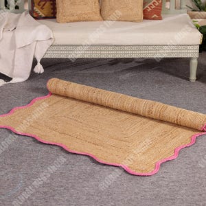 May include: A rolled-up, rectangular jute rug with a scalloped edge and a bright pink border. The natural-colored rug is partially unrolled, revealing its woven texture. The rug is on a gray surface.