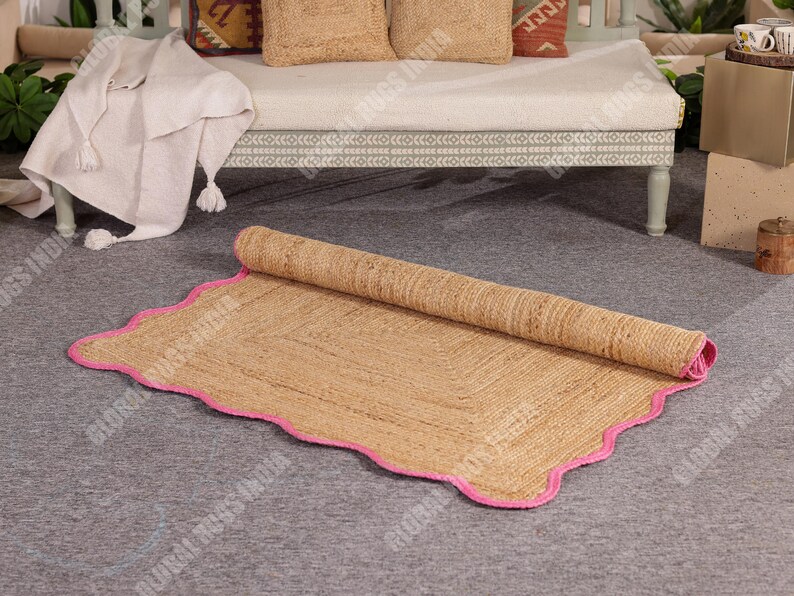 May include: A rolled-up, rectangular jute rug with a scalloped edge and a bright pink border. The natural-colored rug is partially unrolled, revealing its woven texture. The rug is on a gray surface.