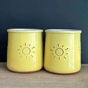 May include: Two cylindrical ceramic mugs with a glossy yellow glaze and a white interior. Each mug features a debossed sun icon on the front, consisting of a central circle surrounded by short, radiating lines. The base of each mug is unglazed terracotta.