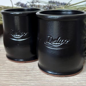 May include: Two cylindrical black ceramic cups with a glossy finish and a slightly flared rim. Each cup features the word Deluxe embossed in a cursive script on the front. The base of each cup reveals a natural terracotta-coloured clay rim.