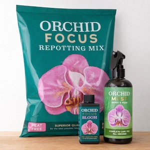 May include: A teal bag of Orchid Focus Repotting Mix featuring a pink orchid graphic, a small black bottle of Orchid Focus Bloom fertiliser, and a dark brown spray bottle of Orchid Focus Myst Spray & Feed with a pink orchid illustration.
