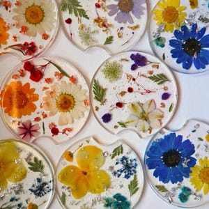 May include: A collection of clear resin car coasters, each embedded with colorful dried flowers and metallic flakes. The coasters feature a variety of floral arrangements, including daisies, and other wildflowers, in shades of yellow, orange, blue, and white.