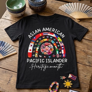 Può includere: T-shirt nera con un design arcobaleno con bandiere e fiori, con la scritta "ASIAN AMERICAN PACIFIC ISLANDER Heritage month". Spilli decorativi con bandiere e disegni floreali sono sparsi.
