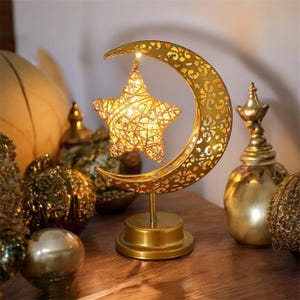 Op de afbeelding: Een decoratieve goudkleurige lamp met een maan en ster ontwerp. De maan heeft ingewikkelde uitgesneden patronen en de ster is gemaakt van geweven materiaal. De lamp wordt verlicht met een warm licht, wat een gezellige sfeer creëert. De basis is rond.