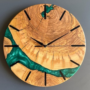 May include: A round wooden clock with a natural wood grain pattern and a vibrant green epoxy resin river design. The clock features black hour markers and clock hands. The clock's diameter is approximately 12 inches.