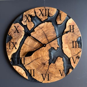 May include: A round wooden wall clock with a natural wood grain pattern and black resin accents. The clock features black Roman numerals and black clock hands. The clock's diameter is approximately 18 inches.