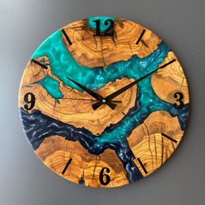Könnte beinhalten: Eine runde Wanduhr aus Holz mit einem einzigartigen Design. Das Zifferblatt besteht aus Naturholz mit einem türkisfarbenen und marineblauen Harzfluss-Design. Schwarze Zeiger und Ziffern zeigen die Zeit an. Die Uhr hat einen Durchmesser von ca. 30 cm.
