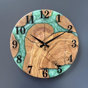 May include: Circular wooden wall clock featuring a natural wood grain design and a turquoise resin inlay. Black numerals mark the hours, and black hands indicate the time. The clock has a modern, rustic aesthetic.