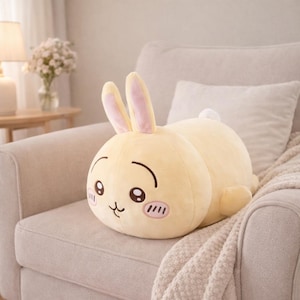 Könnte beinhalten: Ein gelbes Plüsch-Kaninchenkissen mit einem lächelnden Gesicht, rosa Wangen und langen Ohren mit rosa Innenseiten. Das Stofftier liegt auf einem beigen Sofa mit einer weißen Decke.