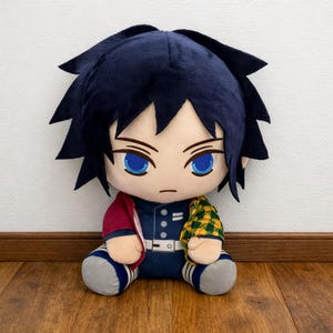 May include: Plush doll of a character with dark blue hair and blue eyes. The doll wears a dark blue uniform with a red jacket and a yellow and green patterned sleeve. The doll is sitting on a wooden floor.
