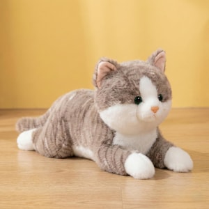 Könnte beinhalten: Ein Stofftier Katze in entspannter Pose. Die Katze hat grau-weiß gestreiftes Fell, weiße Pfoten und einen weißen Fleck im Gesicht. Sie hat grüne Augen und eine rosa Nase. Der Hintergrund ist einfarbig gelb.