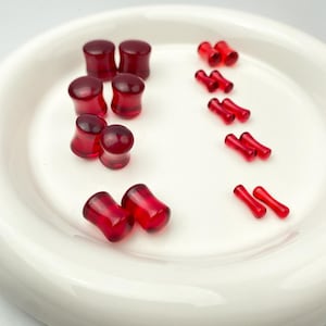 Puede incluir: Una colección de tapones y expansores para oídos rojos y translúcidos dispuestos sobre un plato blanco. Los tapones son cilíndricos con extremos acampanados, mientras que los expansores son alargados y cónicos. Los artículos están espaciados uniformemente.