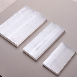 Puede incluir: Tres losas rectangulares de cristal de selenita blanco translúcido dispuestas en orden de tamaño descendente. Cada pieza presenta una textura lineal y fibrosa distintiva que recorre verticalmente la superficie, creando un aspecto nacarado brillante.