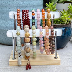 Peut inclure: Un présentoir à bijoux en bois avec plusieurs bracelets de perles de différentes couleurs : rouge, marron, rose, orange et vert. Le présentoir comporte deux barres horizontales blanches soutenues par des poteaux en métal argenté.