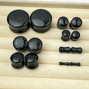 Könnte beinhalten: Eine Sammlung schwarzer Ohrplugs und Tunnel mit dezenten grünen Glitzerpartikeln. Das Set enthält verschiedene Größen von runden Plugs und kleineren zylindrischen Tunneln, die alle eine glatte, polierte Oberfläche und einen dunklen Glanz aufweisen.