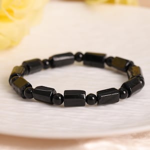 May include: A stretch bracelet featuring alternating black cylindrical beads and smaller black spherical beads. The polished black beads are arranged in a repeating pattern to form a circular band, creating a uniform and dark aesthetic.