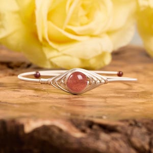 May include: A wire-wrapped bracelet featuring a central round, polished, reddish-pink gemstone. The silver-toned metal wire is coiled intricately around the stone and extends into a thin, smooth band with small dark beads at the ends.
