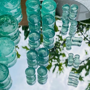 May include: An assortment of teal-colored glass plugs and tunnels are arranged on a reflective surface. The plugs vary in shape, including stacked cylinders and rounded ends. The tunnels have a flared design. The objects are displayed against a backdrop of greenery.