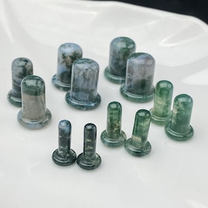May include: A selection of green and grey stone plugs with a flared base and cylindrical top. The plugs are of varying sizes and are arranged on a white surface. The stone has a natural, mottled appearance.