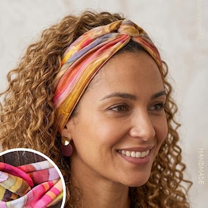 Könnte beinhalten: Ein handgefertigtes Stirnband mit einem Knoten-Design. Das Stirnband zeigt ein lebendiges Muster aus rosa, orange, gelben und lila Streifen. Die Person mit lockigem braunem Haar und goldenen Ohrringen trägt das Accessoire. Das Bild enthält auch eine Nahaufnahme des Stoffes.