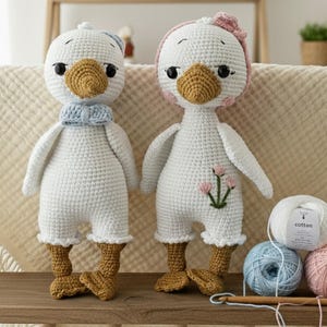 Handmade amigurumi crochet goose dolls (boy and girl), standing on a wooden shelf in a nursery room, accompanied by cotton yarn skeins and a crochet hook. Perfect baby shower gift or unique nursery decor.