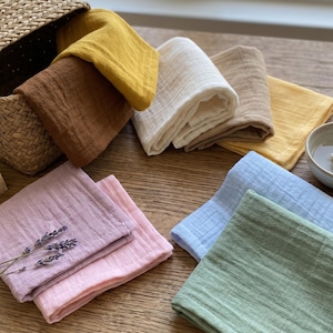 May include: A collection of folded, square washcloths in various colors, including yellow, brown, white, pink, blue, and green. Some cloths are stacked, while others are arranged on a wooden surface. A woven basket and a small bowl are also present.