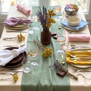 May include: A dining table set with plates, napkins, and silverware, with a light green runner. Wine glasses and a brown vase with dried flowers are present. The table setting includes pink, yellow, and white elements, creating a colourful display.