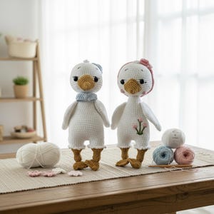 Handmade amigurumi crochet goose dolls displayed on a rustic wooden table with various colored cotton yarn skeins, crochet hooks, and small flower appliques, emphasizing the artisanal craft.