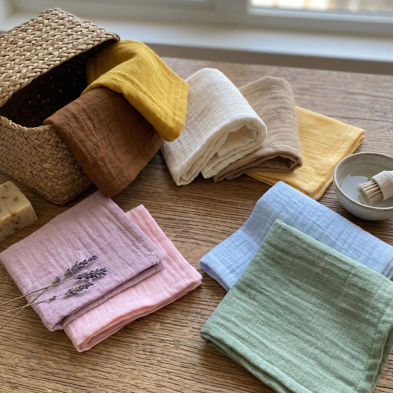 May include: A collection of folded, square washcloths in various colors, including yellow, brown, white, pink, blue, and green. Some cloths are stacked, while others are arranged on a wooden surface. A woven basket and a small bowl are also present.