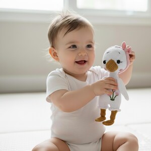 A joyful baby, sitting on a soft play mat, happily hugging a pink-bonneted, flower-embroidered amigurumi goose doll. A perfect soft toy for toddlers and a cherished kids gift.