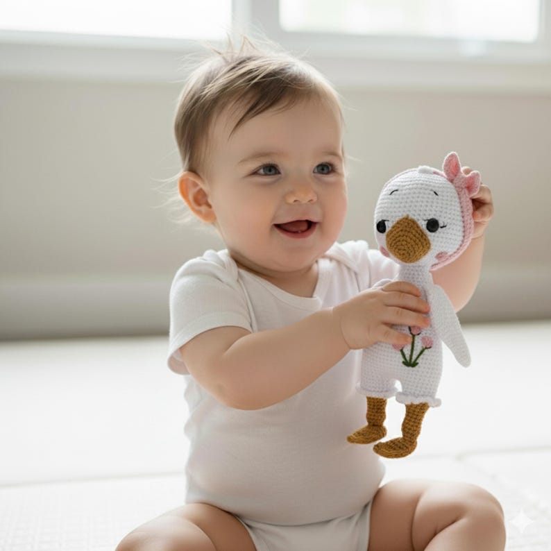 A joyful baby, sitting on a soft play mat, happily hugging a pink-bonneted, flower-embroidered amigurumi goose doll. A perfect soft toy for toddlers and a cherished kids gift.