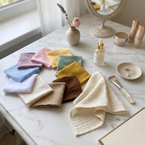 Pastel and earth tone muslin cleansing cloths arranged on a marble vanity with skincare serums. Gentle exfoliating facial squares, reusable organic makeup wipes, luxury eco-friendly beauty routine gift, soft cotton hanky.